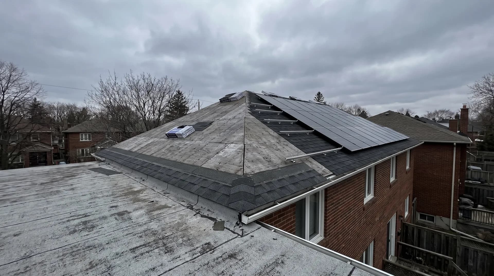A brick house roof photographed from next door, half of it stripped back to grey plywood decking with a first course of new shingles along the eave, the other half already carrying black solar modules.